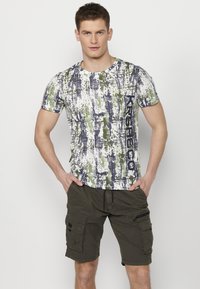 T-shirt s kratkim rukavima s bijelim, zelenim i modrim apstraktnim uzorkom; nosi se uz maslinaste cargo kratke hlače s bočnim džepovima.