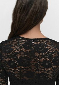 Zwart kanten top met een bloemenpatroon, ronde halslijn en een kleine ronde accent achteraan. Zachte textuur met doorschijnende gebieden.