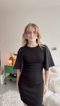 Czarna sukienka z krótkimi falbaniastymi rękawami, dopasowany krój i długość do kolan. Gładki materiał i minimalne zdobienia. Jasne tło z zielonymi meblami.