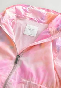 Pink-orange Batik-Zipjacke mit Kapuze und weißem Stoffetikett mit der Aufschrift "NEXT Made in Myanmar" innen am Kragen.