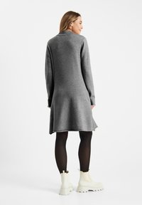 Grijze gebreide jurk met lange mouwen, V-hals en uitlopende zoom, gecombineerd met witte enkellaarsjes en zwarte panty’s. Zachte textuur, casual ontwerp.