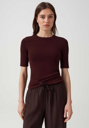 Junge Frau mit glattem braunem Haar, die ein eng anliegendes dunkelburgunderfarbenes Shirt und lockere, strukturierte dunkelbraune Hose trägt, stehend und nach vorne blickend.