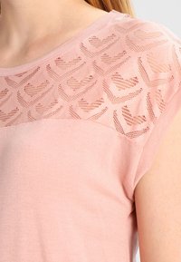 Detailní záběr na světle růžový tílko bez rukávů s texturovaným srdcem a chevron vzorem krajky podél horní části hrudníku a ramenní oblasti.