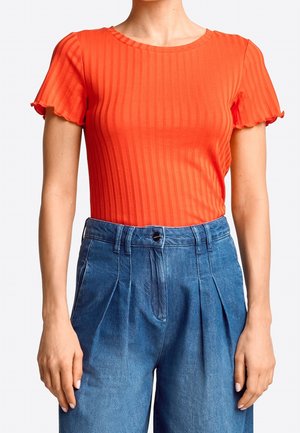 Kobieta w dopasowanej, prążkowanej, pomarańczowej bluzce z krótkim rękawem i falbaniastymi krawędziami oraz w granatowych, wysokich, plisowanych dżinsach.