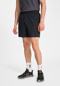 Svarta träningsshorts med elastisk midja, tillverkade av lättviktig tyg. Bärs med svarta sneakers och vita strumpor med ett chevronmönster.