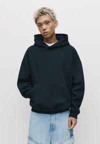 Sötét navy kék, oversize kapucnis pulóver, pamutból készült, elülső zsebbel és húzózsinórral a kapucnin. Világoskék farmer rövidnadrággal párosítva.