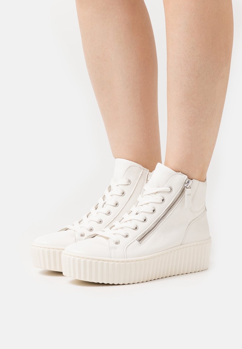 Białe wysokie sneakersy na platformie z bocznym zamkiem i sznurowadłami noszone na nagich nogach na białym tle.