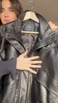 Černá kožená bunda s texturovaným povrchem, zatrhávacími knoflíky a podšívkou. Visí na dřevěném ramínku na neutrálním pozadí.