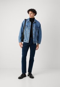 Denim jakna v svetlo modri barvi, črn pulover s turtle-neck, temno modri žametni hlače in črne čevlje. Model drži črno torbico za ramo, stoji ob svetli kulisi.
