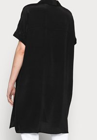 Abito camicia nero a maniche corte con vestibilità rilassata, tessuto morbido, colletto e fessure laterali, caratterizzato da un retro lungo e un orlo dritto.