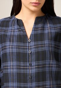 Niebieska i czarna koszula w kratę na guziki, z dekoltem w serek, krótkimi rękawami i pięcioma ciemnymi guzikami z przodu. Miękka faktura tkaniny.