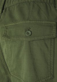 Olive green cargo shorts feature a front pocket with a flap, secured by a single button, made from textured fabric with visible stitching.