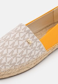 Primo piano di una scarpa espadrilla beige e gialla con suola in juta intrecciata e motivo ripetuto "MK" nella parte superiore.