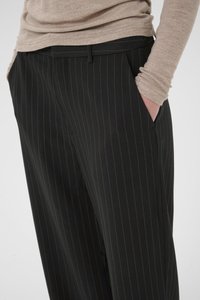 Sorte pinstripede bukser laget av glatt stoff, med skrå frontlommer og tilpasset passform. Pinstripene er i en kontrastfarge.
