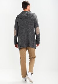 Cárdigan de punto en gris carbón con capucha, con parches en los codos, un dobladillo trasero largo, combinado con pantalones beiges y zapatillas blancas.