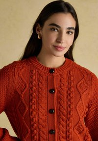 Oranžs adīts kardigans ar diegu rakstiem, apaļu kakla izgriezumu un četriem melniem pogām gar priekšpusi. Faktūras audums ar siltu izskatu.
