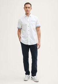 Korte mouwen, wit overhemd met knoopsluiting en blauwe accenten op de manchetten, gecombineerd met navy broek en witte sneakers. Heeft een borstzak en bruine knopen.