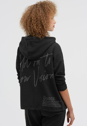 Woman with curly hair wearing a black hoodie with hood down, showing handwritten text and a motivational quote on the back.