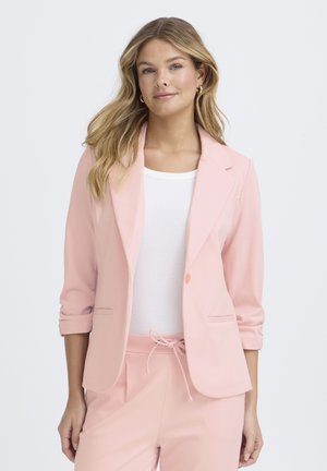 Ženska z valovitimi blond lasmi, ki nosi zlate obročaste uhane, belo majico in svetlo rožnati blazer s pripadajočimi hlačami z vrvico, nežno se smehlja.