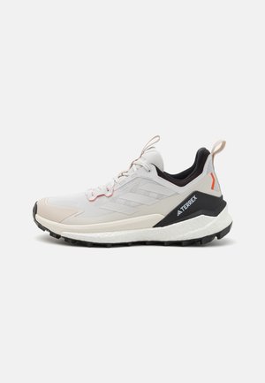 Leichte Wanderschuhe mit einem Obermaterial aus weißem und beigem Mesh, schwarzen Akzenten, roten Details, einer strukturierten Sohle und auffälliger Markenkennung an der Ferse.