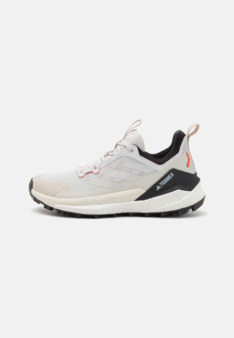 Shoe de caminhada leve com um cabedal em malha branca e bege, detalhes em preto, pormenores em vermelho, sola texturizada e marca em destaque no calcanhar.
