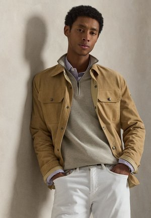 Junger Mann lehnt an der Wand, trägt eine hellbraune Jacke, einen beigen Reißverschluss-Pullover, ein gestreiftes Hemd und weiße Hose mit den Händen in den Taschen.