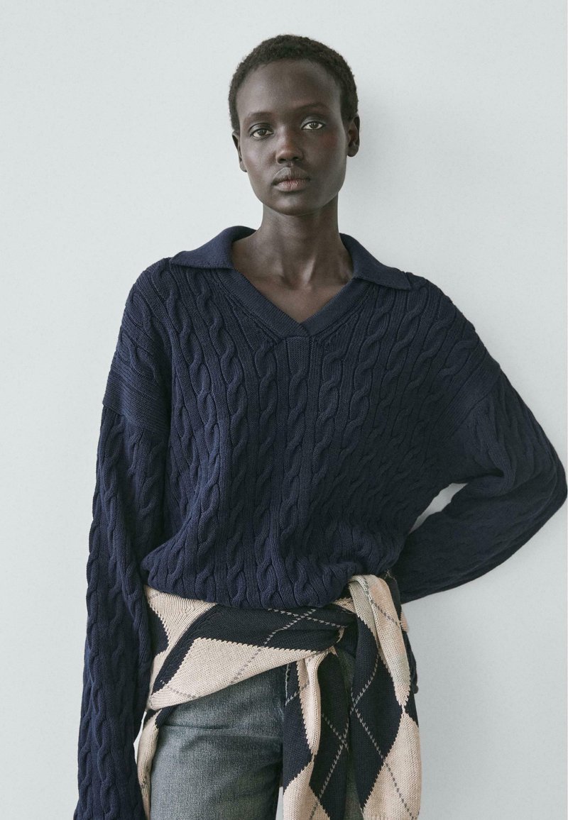 Person med kort hår iført en marineblå kabelstrikket sweater og et sort-beige mønstret tøj bundet omkring livet.