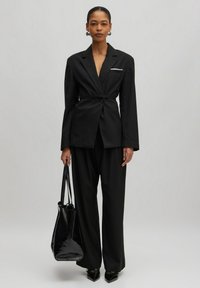 Schwarzer maßgeschneiderter Anzug mit tailliertem Gürtel, strukturierten Revers und einem weißen Taschendetail. Kombiniert mit einer glänzenden schwarzen Tasche und spitzen Absätzen.