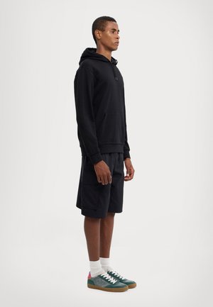 Felpa nera con cappuccio e tasca frontale, abbinata a pantaloni corti neri. Il modello indossa sneaker verdi e grigie con dettagli rosa e calzini bianchi.