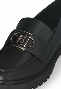 Mocassino in pelle nera con suola spessa, dotato di una fibbia con logo dorato su una larga fascia. Texture liscia con cuciture dettagliate.