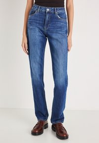 Mėlyni tiesaus kirpimo džinsai su aukštu liemeniu, klasikiniu penkių kišenių dizainu ir subtiliu išblukimu. Derinami su rudos spalvos odiniais slip-on batais.