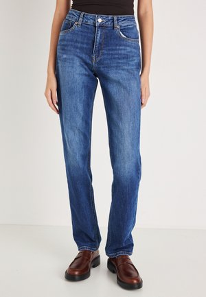 Jean droit bleu avec taille haute, design classique à cinq poches et légère décoloration. Associé à des chaussures en cuir marron à enfiler.