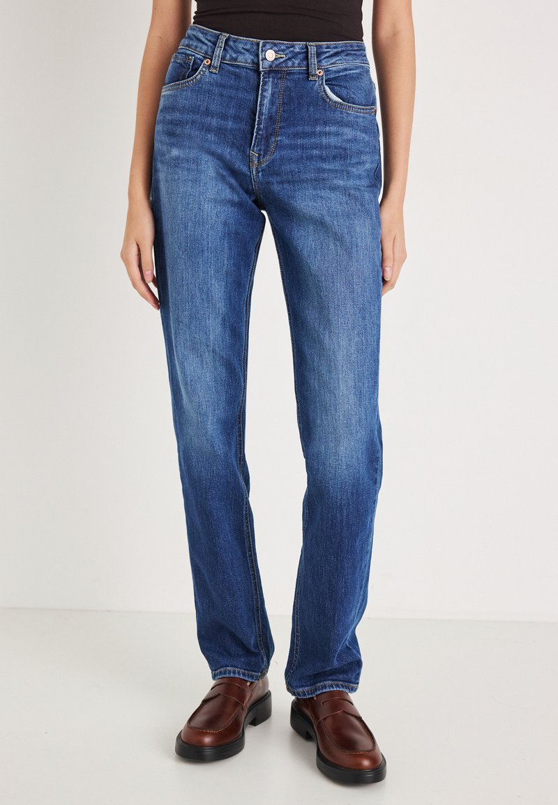 Mėlyni tiesaus kirpimo džinsai su aukštu liemeniu, klasikiniu penkių kišenių dizainu ir subtiliu išblukimu. Derinami su rudos spalvos odiniais slip-on batais.