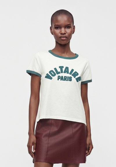 Γυναίκα που φοράει λευκό T-shirt με πράσινο περίγραμμα και κείμενο "Voltaire Paris", συνδυασμένο με σκουρόχρωμη κόκκινη δερμάτινη φούστα σε απλό φόντο.