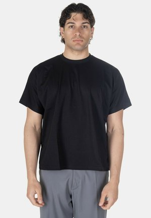Giovane uomo con capelli ricci scuri indossa una maglietta nera semplice e pantaloni grigi, in piedi di fronte a uno sfondo chiaro.