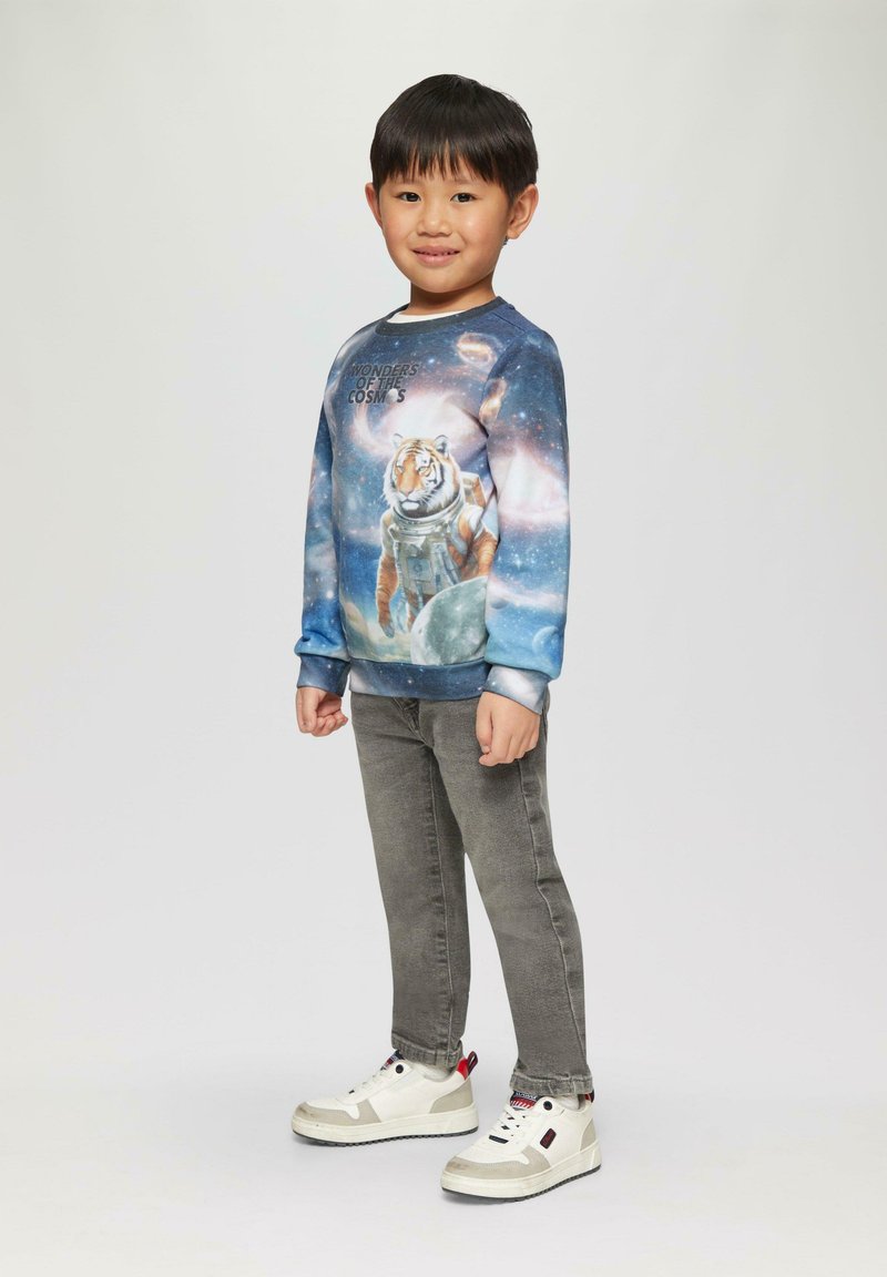 Blauwe sweatshirt met een tijger in een astronautenpak, versierd met een galaxiepatroon. Grijze jeans en witte sneakers met rode accenten maken de outfit compleet.