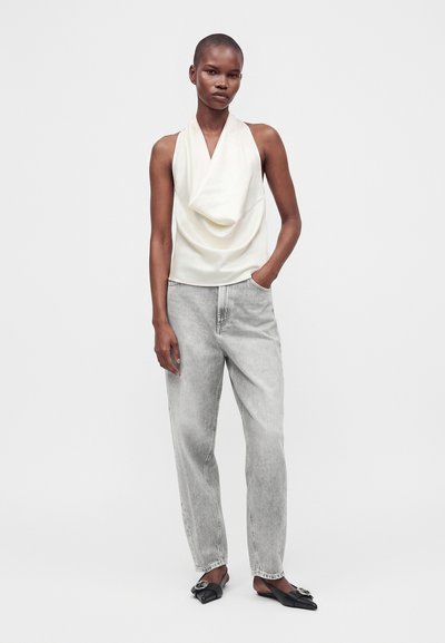 Jeune personne portant un haut blanc sans manches drapé, un jean gris clair et des ballerines noires pointues, debout avec une main dans la poche.