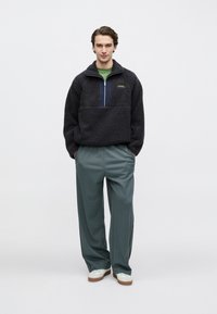 Φλις πουλόβερ σε σκούρο γκρι με φερμουάρ τέταρτου μήκους, με αντίθετη μπλε επιφάνεια, σε συνδυασμό με φαρδιά πράσινα παντελόνια και λευκά αθλητικά παπούτσια.