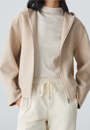 Kvinde iført beige hoodie med lynlås over en lys, mønstret ribstrikket top og cremelignende bukser med snøre, hænderne i lommerne.