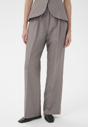 Kobieta ubrana w luźne, szare, dopasowane spodnie z przodu z zakładkami i czarne buty z kwadratowymi noskami, stojąca na jednolitym tle.