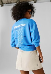 Γυναίκα με σγουρά μαλλιά που φοράει γυαλιά ηλίου, μπλε φούτερ με την επιγραφή «World Tour» και λίστα παγκόσμιων πόλεων, καθώς και μπεζ φούστα, στέκεται μπροστά σε ένα απλό φόντο.