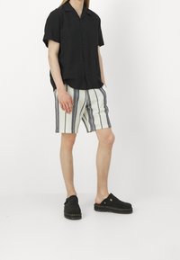 Osoba nosząca czarną koszulę z krótkim rękawem zapinaną na guziki, białe i granatowe szorty w paski oraz czarne buty wsuwane na białym tle.