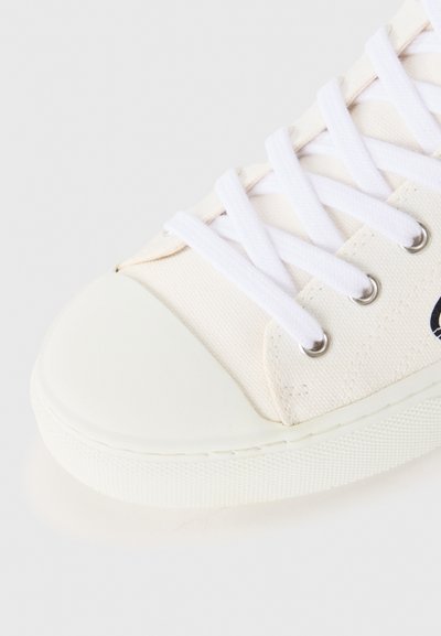 Canvas sneaker in off-white with a rubber toe cap, white laces, silver eyelets, and a textured sole. Minimalist design with no prominent patterns.