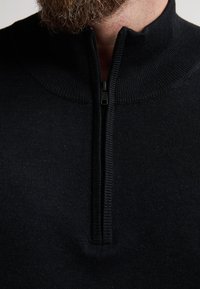 Schwarzer Zip-Up-Pullover mit geripptem Kragen und strukturiertem Stoff. Verfügt über einen Reißverschluss in Viertellänge vorne. Nahaufnahme des Ausschnitts.