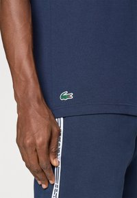 Ναυτικό κοντομάνικο μπλουζάκι από βαμβάκι με πράσινο και κόκκινο λογότυπο κροκόδειλου στο στρίφωμα, συνδυασμένο με σκούρες μπλε παντελόνες με λευκές πλαϊνές ρίγες.