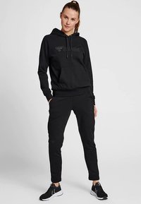 Czarna bluza z kapturem i kieszenią typu kangur, ze ściągaczem przy kapturze, w połączeniu z czarnymi, dopasowanymi dresami. Oba elementy mają miękką teksturę materiału.