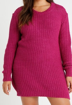 Vrouw draagt een langarmige, gestructureerde gebreide magenta trui-jurk, staand met ontspannen armen langs haar zij.