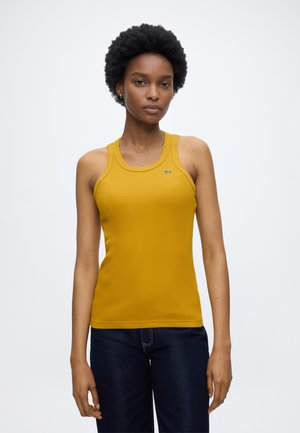 Femme aux cheveux courts et bouclés portant un haut sans manches côtelé couleur moutarde et un jean foncé, se tenant devant un fond clair uni.