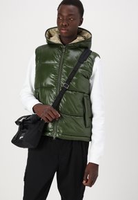 Groene, glanzende puffervest met capuchon en beige voering, met een ritssluiting aan de voorkant en twee zijzakken, gedragen over een wit sweatshirt.