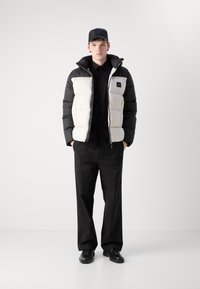 Pufferjakke med sorte og cremede paneler, med hætte og lynlås. En tætsiddende sort skjorte og sorte vidde bukser fuldender outfittet.